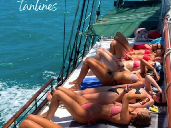 People sunbathing in bikinis on a sailboat with ocean and mountains in the background.