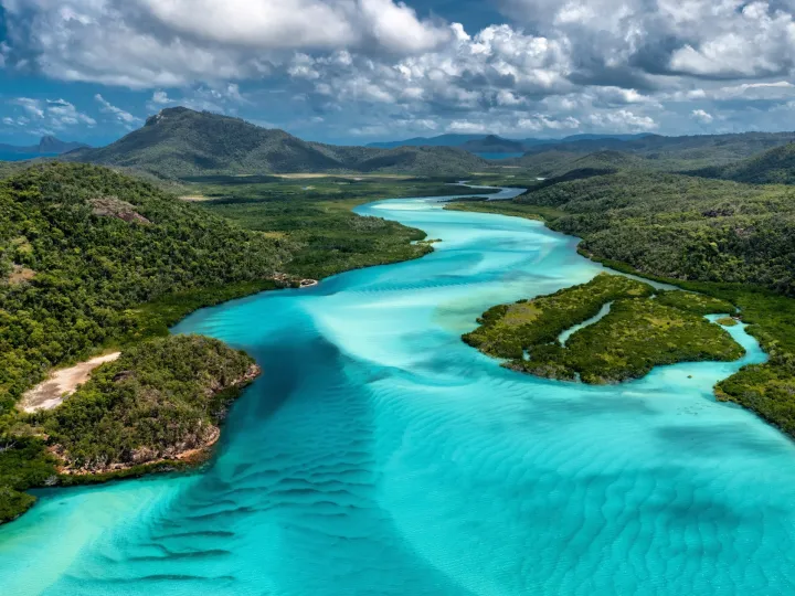 Aerial view of a winding turquoise river through lush green mountains under a partly cloudy sky.